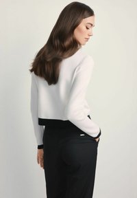 Biały, krótki sweter z czarnym prążkowanym wykończeniem, z długimi rękawami. Modelka w czarnych spodniach, stoi z rękami w kieszeniach. Płaskie tło.