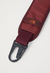 Burgundi színű, textúrált felületű pánt, fekete műanyag csattal és a The North Face kis narancssárga logójával.