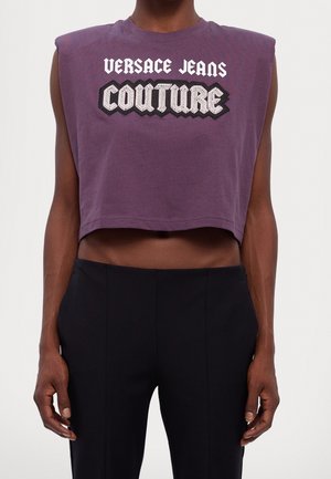 Ærmeløs lilla cropped top med teksten "VERSACE JEANS COUTURE" båret sammen med højtaljede sorte bukser på en stående person.