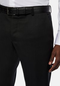 Črne krojene hlače z gladko teksturo, ki vključujejo črn usnjen pas s kvadratno sponko. Delno vidna bela srajca.