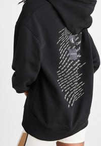 Person som bär en svart hoodie med vit textdesign som upprepas i form av en diamant med "Calvin Klein Jeans" på baksidan.