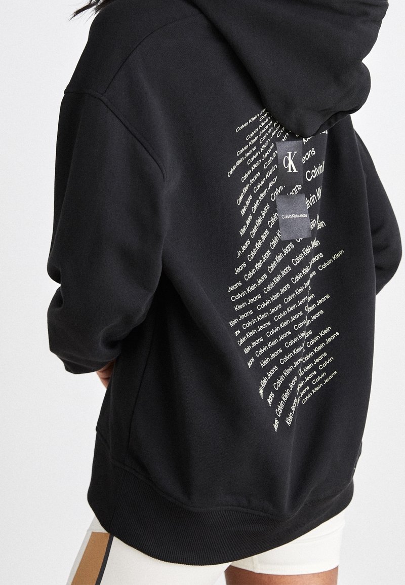 Person som bär en svart hoodie med vit textdesign som upprepas i form av en diamant med "Calvin Klein Jeans" på baksidan.