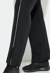 Czarne sportowe spodnie o luźnym kroju, z białym paskiem po bokach, rozcięciem przy hemie, wykonane z miękkiego materiału, w połączeniu z czarnymi trampkami.