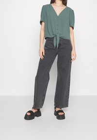 Haut vert à manches courtes avec col en V et lien au bas, associé à un jean noir taille haute à jambes larges et des sandales noires épaisses.