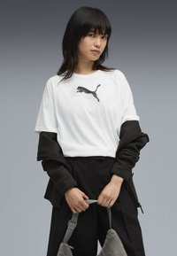 Hvid T-shirt med sort Puma-logo, båret under en mørk jakke. Modellen holder en grå taske med en blød, pelsagtig tekstur. Sorte bukser fuldender looket.