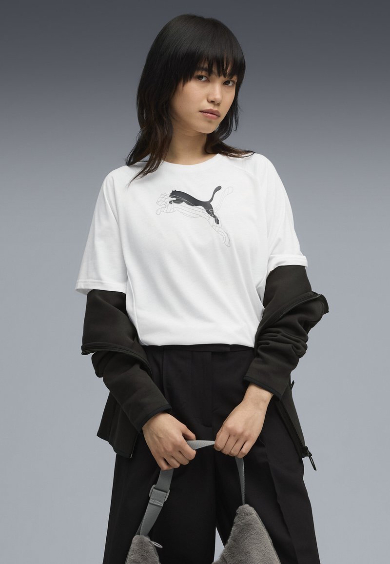 Biała koszulka z czarnym logo Puma, noszona pod ciemną kurtką. Model trzyma szary plecak o miękkiej, futrzastej teksturze. Czarne spodnie dopełniają całość.