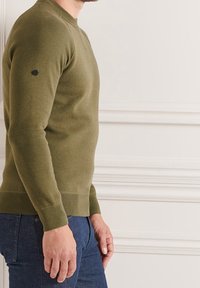 Pull à col rond couleur vert olive, tissu tricoté, poignets et ourlet côtelés, accent sur la manche gauche, porté avec un jean bleu.