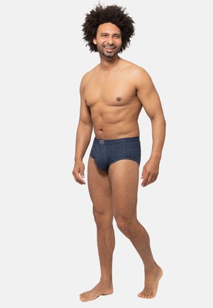 Dunkelblaue gemusterte Slips mit einem strukturierten Design. Der Bund hat ein weiches elastisches Band. Das Model steht barfuß und lächelt.