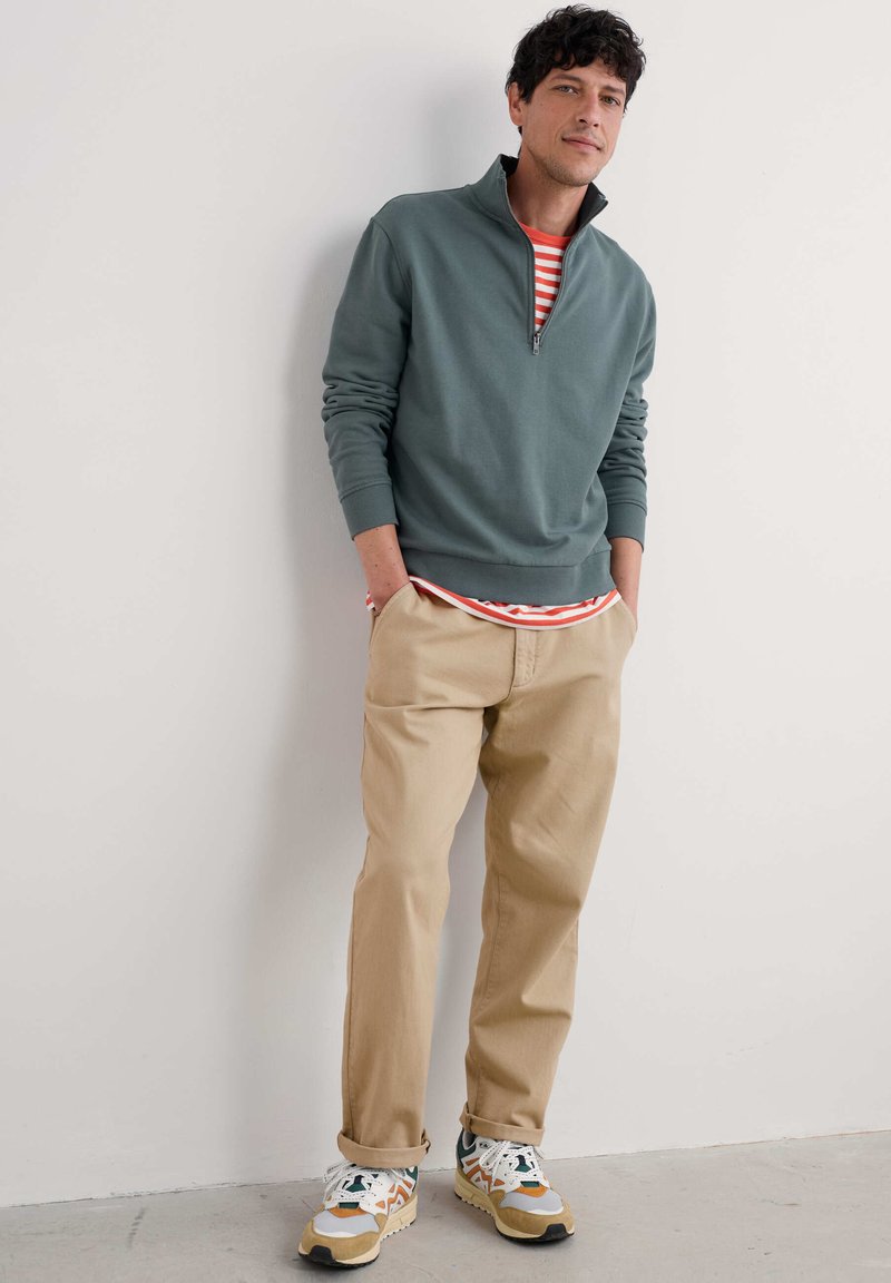Sweatshirt mit Quarter-Zip aus gedämpftem grünem Stoff, übereinander getragen mit einem rot gestreiften Hemd, kombiniert mit beigen Hosen und mehrfarbigen Turnschuhen.
