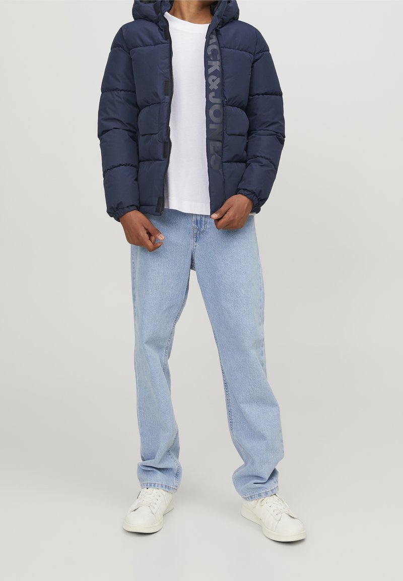 Laivaston sininen puffatakki hupparilla, jossa on näkyvä logo, yhdistettynä vaaleansinisiin farkkuihin ja valkoisiin lenkkareihin. Silkkinen, pehmustettu pinta.