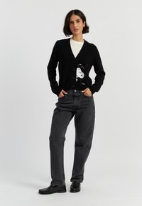 Cardigan nero con un design di Snoopy sul davanti, caratterizzato da scollo a V, chiusura con bottoni e polsini a costine. Indossato sopra una camicia bianca con jeans grigi.