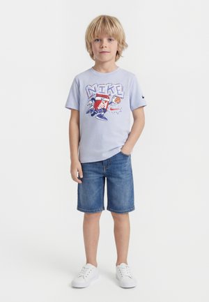 Blond boy wearing a light purple Nike graphic t-shirt, denim shorts, and white sneakers, standing with one hand in pocket.