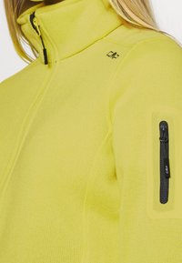 Gekürzte gelbe Fleece-Jacke mit hohem Kragen, Reißverschluss vorne und Seitentasche mit schwarzem Reißverschluss sowie Logodetail auf der Schulter.