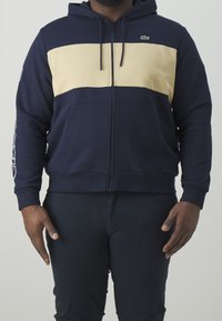 Felpa con zip di colore blu navy con una striscia orizzontale beige. Presenta una tasca a marsupio e un cappuccio con cordino, con un logo sul petto.
