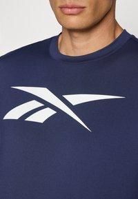 Námornícky modré športové tričko vyrobené z hladkej tkaniny, s výrazným bielym logom na hrudi s ostrými tvarmi. Okrúhly výstrih.