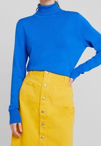 Sininen korkeakauluksinen villapaita, jossa on pitkät hihat, yhdistettynä keltaiseen farkkuhameeseen, jossa on etutaskut ja hopeiset nepparit.