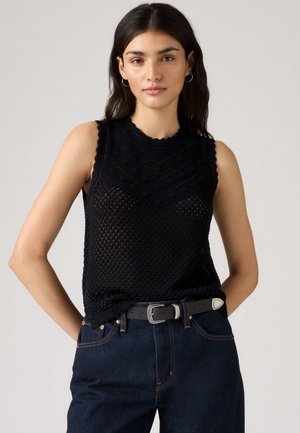 Kvinde iført ærmeløs sort striktop med zigzag mønster, mørkeblå jeans og sort bælte med sølvspænde, stående foran en ensfarvet baggrund.