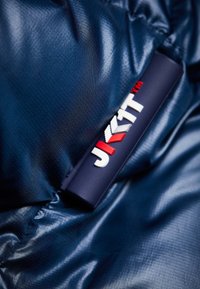 Laivastonsinisen toppatakin materiaali, jossa on sileä, teksturoitu viimeistely. Varustettu mustalla merkillä, jossa on valkoinen ja punainen "JKIT"-brändäys.