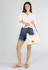 Camisa de botones rosa claro, pantalones cortos de mezclilla, sandalias marrones y un bolso de lona beige con detalle de logotipo y acentos de cuero.