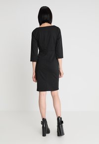 Vrouw staat met de rug naar de toeschouwer gekeerd, gekleed in een zwarte jurk tot de knie met driekwart mouwen en zwarte hoge enkellaarzen.