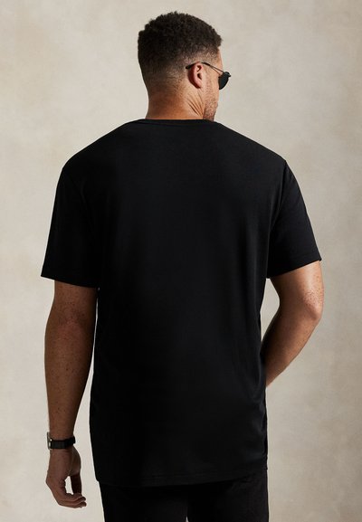 Mann iført en ensfarget svart t-skjorte med korte ermer og solbriller, vist bakfra mot en ensfarget bakgrunn.