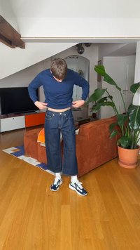 Άνδρας που προσαρμόζει ένα μπλε σκούρο πουλόβερ στο σαλόνι με πορτοκαλί καναπέ, μεγάλο γλαστράκι, ξύλινο πάτωμα και τηλεόραση στο φόντο.