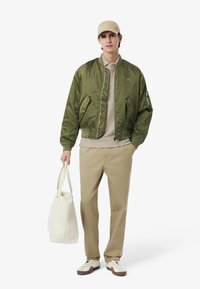 Olivgrön bomberjacka, beige skjorta, khakifärgade byxor och vita sneakers. Modellen håller en vit totebag. Enkel design med minimala accenter.