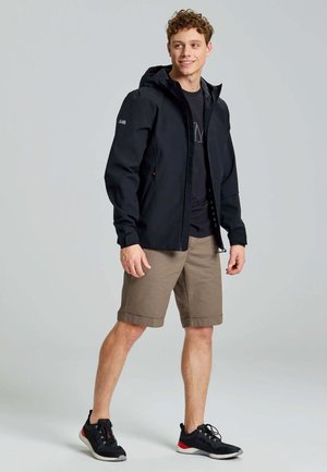 Zwarte waterdichte jas met een capuchon, ritssluiting aan de voorkant en zijzakken, gedragen over een zwart T-shirt, in combinatie met beige shorts en zwarte sneakers.