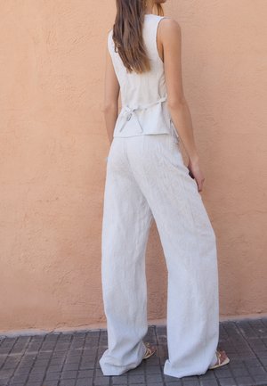 Débardeur gris clair sans manches avec un détail de nœud à l'arrière, associé à un pantalon large à motifs et à coupe décontractée, le tout devant un mur orange texturé.