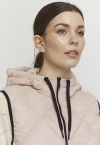 Šviesiai rožinė d quilted liemenė su gobtuvu, su užtrauktuku priekyje ir juodomis nuleidimo virvelėmis, papuošta juodais rankovių išpjovimais ir tekstūruotu megztu sluoksniu po ja.