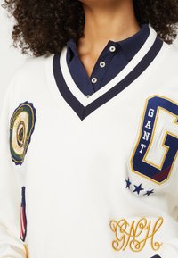 Pull blanc avec un col marine, orné de patchs brodés comprenant un logo, une lettre "G" et des coutures décoratives en or.