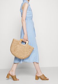 Torbka z plecionego słomy z drewnianym uchwytem, jasnoniebieska sukienka z denimu oraz beżowe, ażurowe buty wsuwane na obcasie w kształcie słupka.