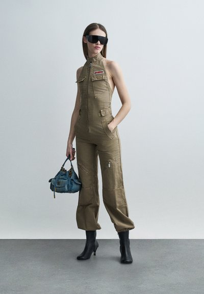 Femme portant une combinaison sans manches kaki avec une fermeture éclair frontale et des écussons, des bottines noires, de grandes lunettes de soleil noires, tenant un sac à main bleu.