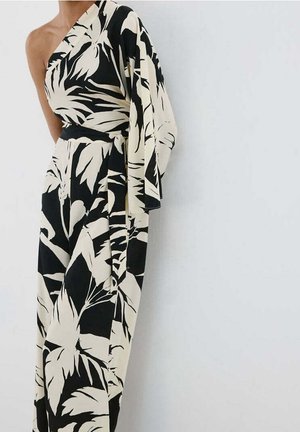 Kvinde iført en sort og hvid one-shoulder jumpsuit med stort bladmønster og vide ben mod en ensfarvet hvid baggrund.