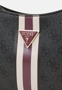 Mustakana tekstuuriga käekott, millel on kolmnurkne Guess USA logo burgundi taustal, mille külgedel on vertikaalsed triibud valge ja burgundi värvuses.