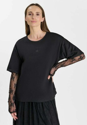 Frau mit langen braunen Haaren trägt ein lockeres schwarzes T-Shirt über langen schwarzen Spitzenärmeln und einem schwarzen Spitzenrock vor einem schlichten weißen Hintergrund.