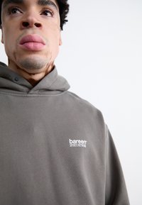 Grå hoodie med en forsigtig lomme og en snørret hætte, med "bareen SPORTS NETWORK" trykt i hvidt. Glat stofstruktur.