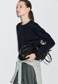 Maglione blu navy con collo rotondo, lavorazione a maglia texturizzata. Il modello tiene in mano una borsa a tracolla in pelle nera con finitura liscia e manici lunghi. Pantaloni in lino grigi.