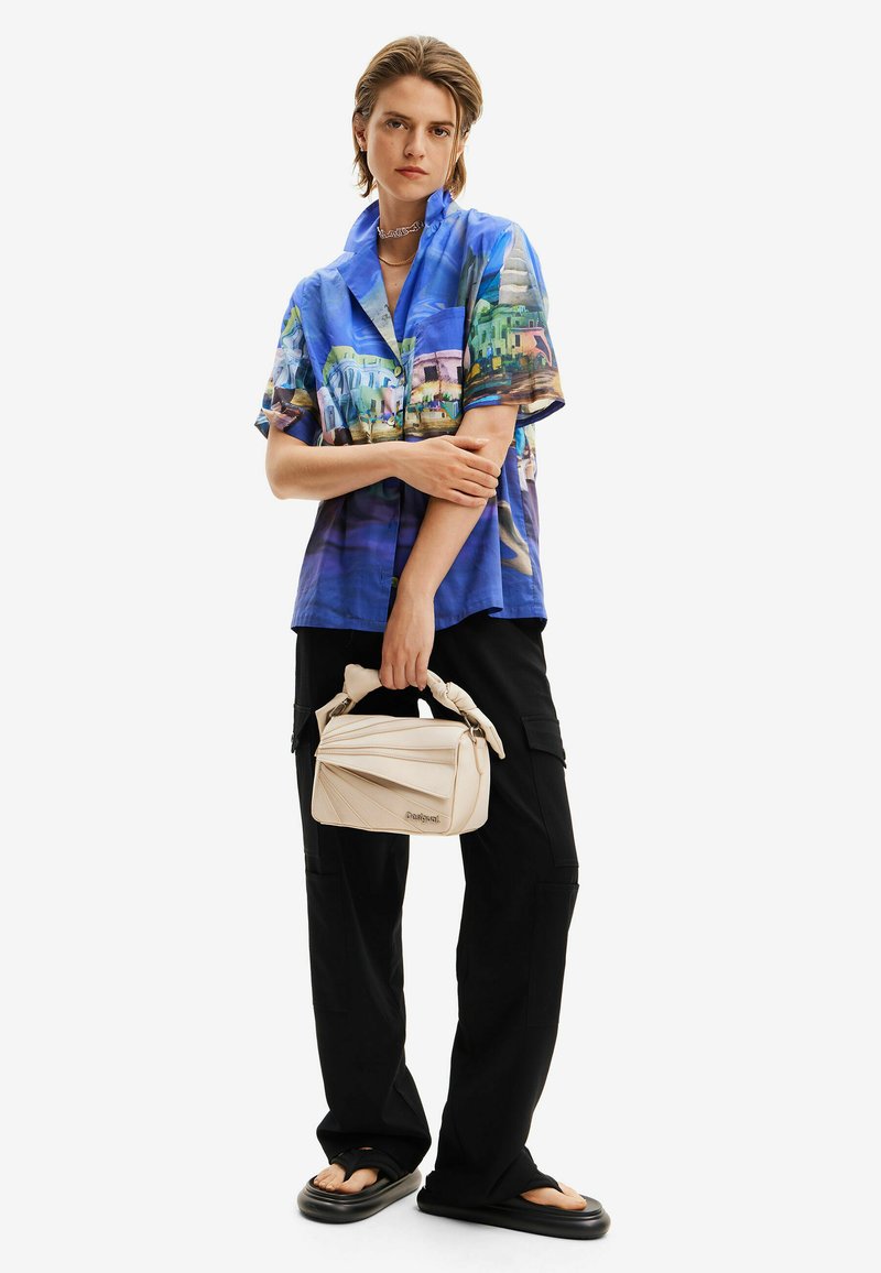 Kurzärmliges, blau gemustertes Hemd mit landschaftlicher Kunst; kombiniert mit schwarzer Hose. Beiger Beutel mit geknotetem Griff und strukturierten Details.