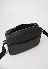 Sort, tekstureret stoftaske med to lynlåslommer, justerbar strop og en glat overflade. Simpelt design med en frontlomme.
