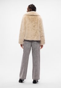 Beige faux fur-jakke med bred krage, parret med grå bredde bukser og kraftige sorte støvler, set bagfra.