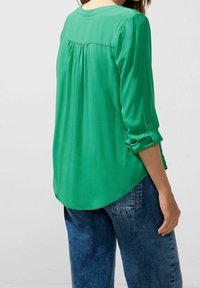 Grön blus med långa ärmar och rund halsringning, bakfållsdetalj och uppvikta manschetter, i kombination med blå denimjeans. Slät textur.