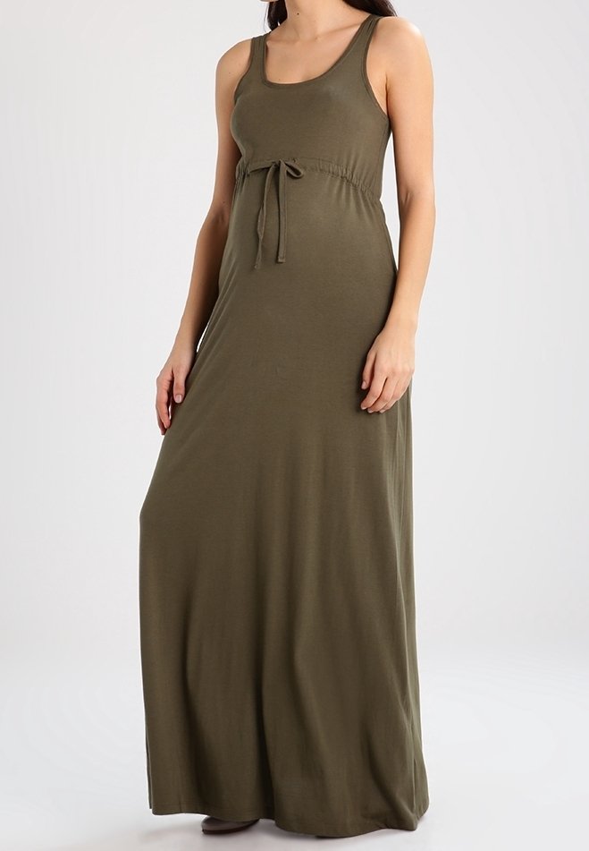Femme portant une robe longue sans manches vert olive avec un détail noué à la taille, debout devant un fond uni.