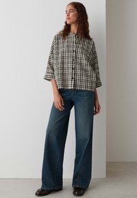 Camicia accorciata a scacchi bianca e nera con collo alto, maniche a tre quarti e bottoni frontali; abbinata a jeans larghi blu.