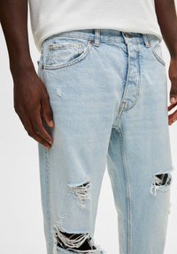 Jeans de mezclilla desgastados en azul claro con desgarros en ambas piernas, un ajuste relajado y costuras en contraste. Presentan bolsillos estándar y un cierre de botón.