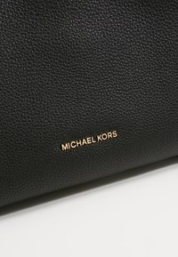 Surface en cuir noir texturé avec petit logo Michael Kors doré centré près du bas sur fond blanc.