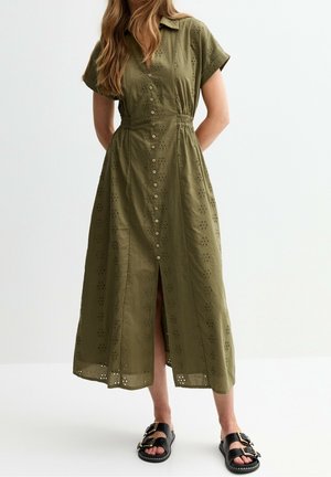Femme portant une robe midi vert olive à motifs floraux ajourés, avec boutons à l'avant, manches courtes, et sandales noires à boucle, debout sur un sol blanc.