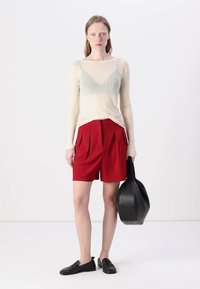 Top long à manches longues en crème, léger et transparent, à côtes, avec des accents teal, associé à des shorts rouges taille haute ; chaussures noires à enfiler, sac oval noir.