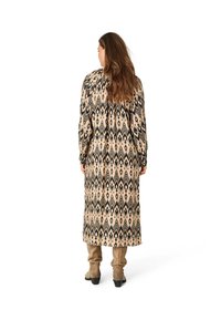 Langes beigefarbenes Kleid mit schwarzem geometrischem Muster, langen Ärmeln und lockerer Passform, kombiniert mit hellbraunen Stiefeln bis zum Knie. Weiche Stofftextur.