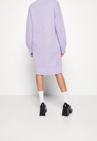 Robe tricot lilas avec un motif texturé, poignets et ourlet côtelés. Associée à des chaussures à talons carrés noires et des chaussettes blanches sous la cheville.